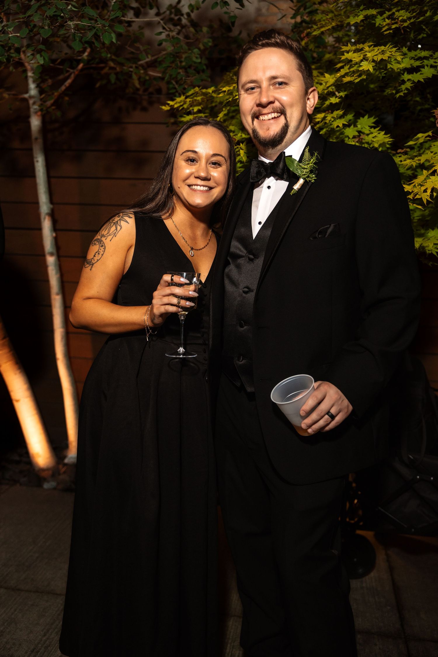 Couple in black formal attire smiling at wedding reception, woman holding wine glass, man in tuxedo with boutonniere.