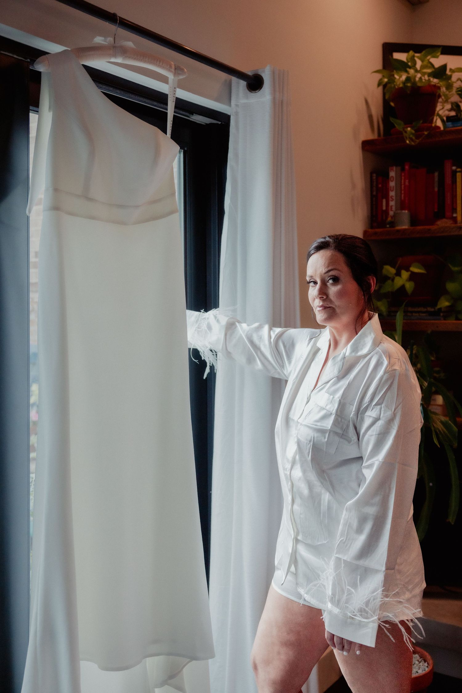 Bride in white robe standing beside elegant strapless wedding gown hanging near bright window.