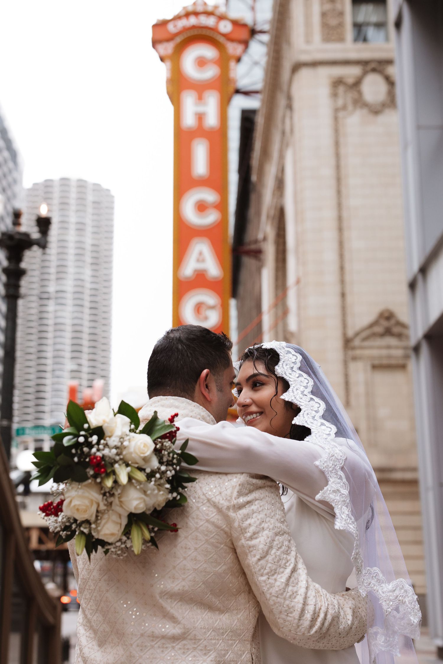 Bride in lace veil smiles over groom's shoulder in front of Chicago Theatre sign.