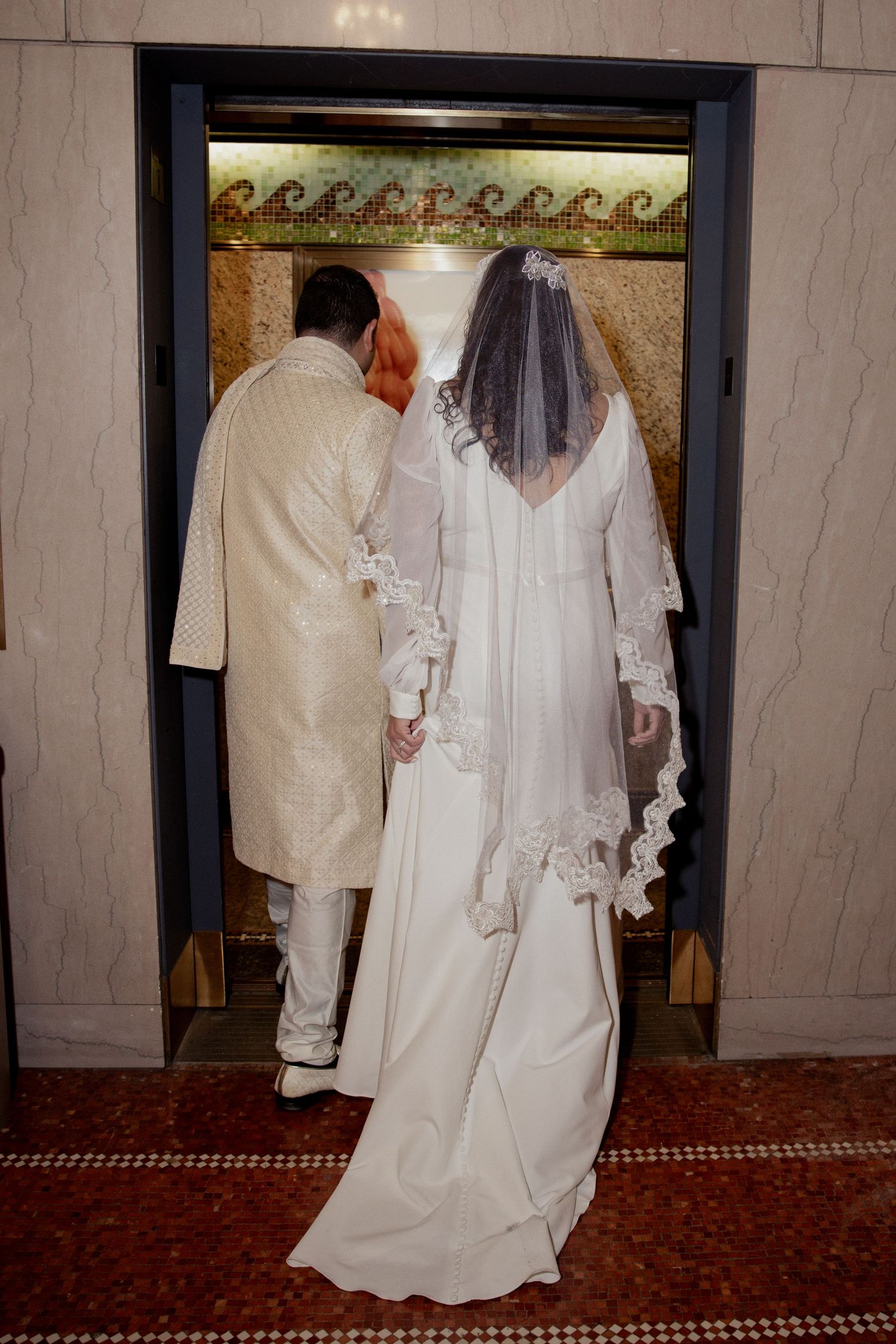Bride in lace veil and groom in sherwani enter a decorative elevator together.