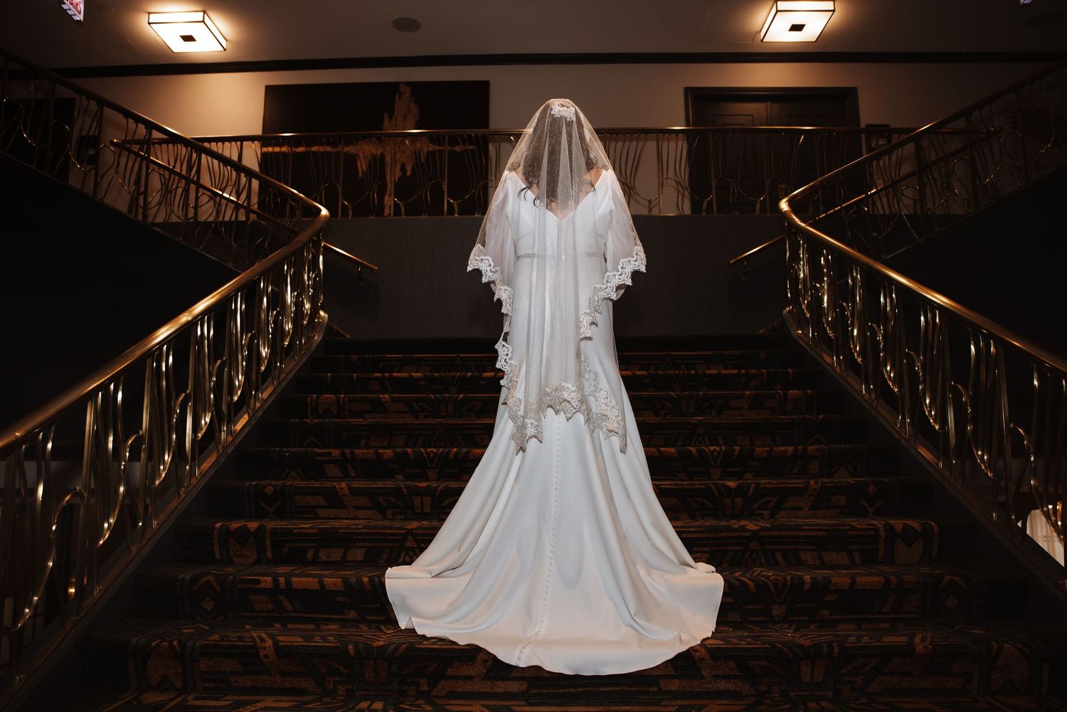 Back view of bride in white wedding gown with lace veil standing on grand staircase with gold railings.