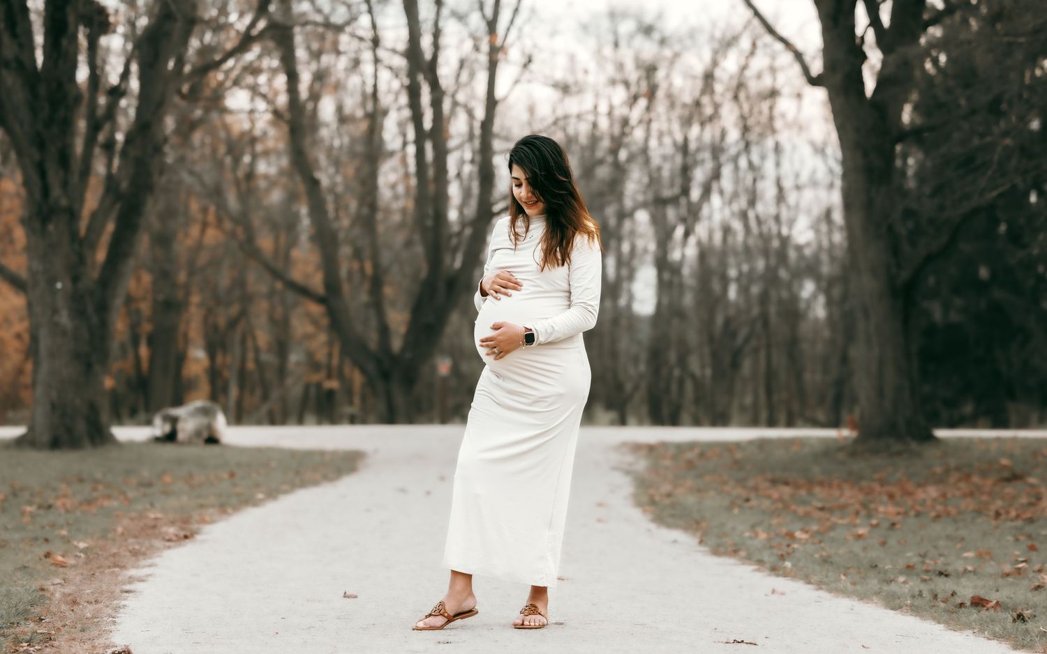 Pregnant woman in white dress standing alone on a park path surrounded by autumn trees.