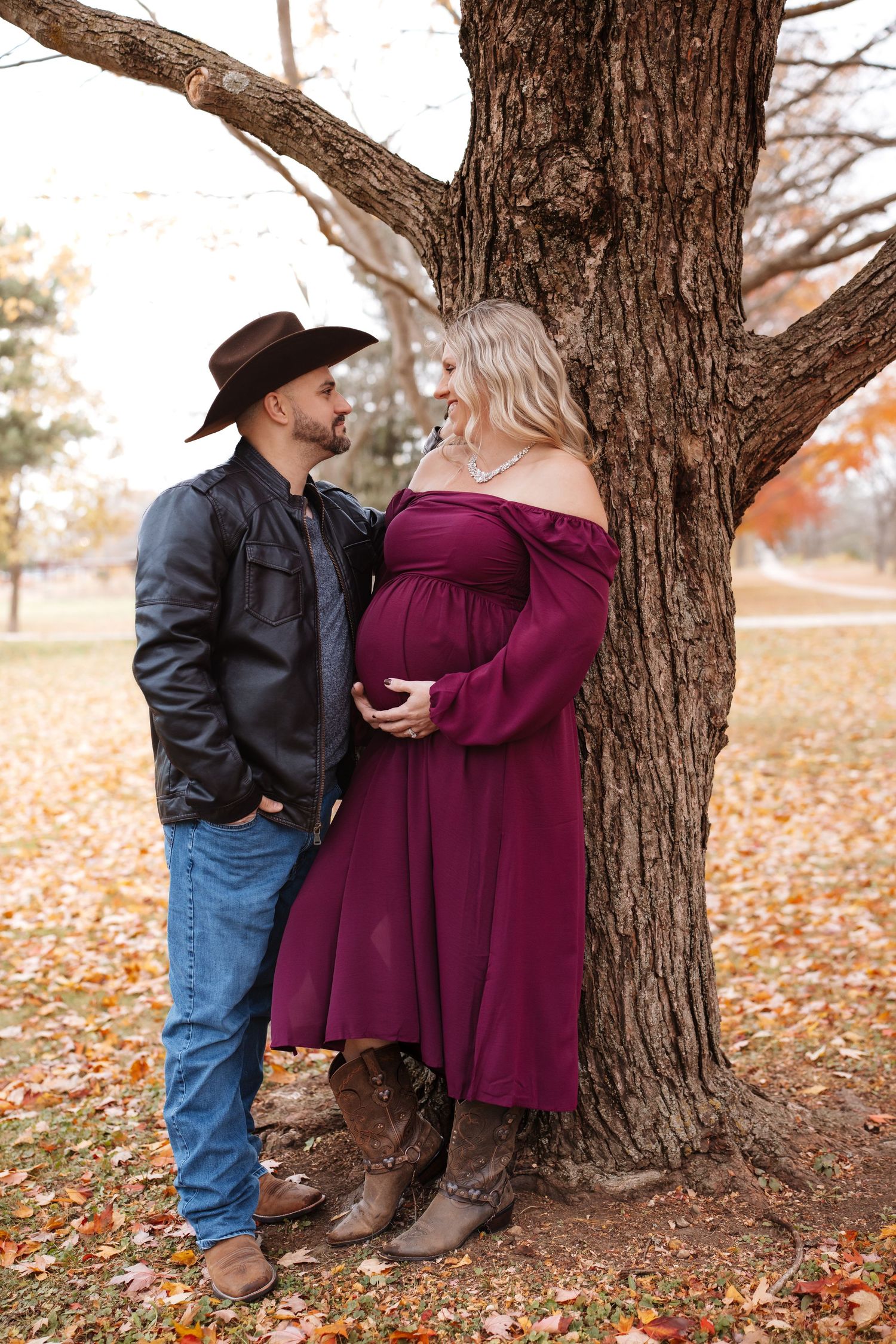 Couple sharing a loving gaze by a tree, man in cowboy hat with pregnant woman in burgundy off-shoulder dress.