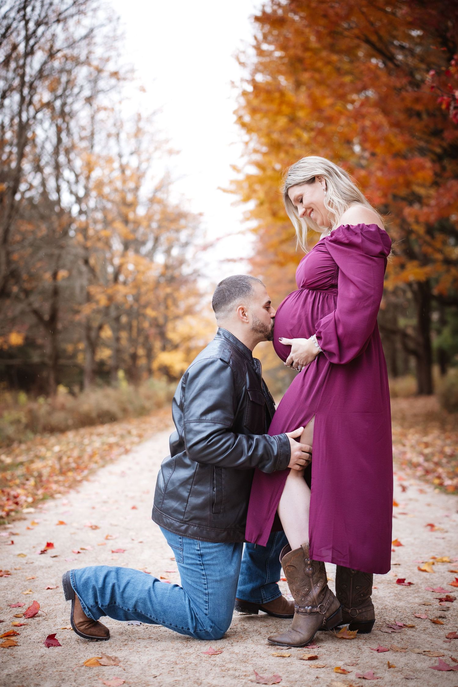 Man kneeling and kissing pregnant woman's belly in a purple dress on a colorful autumn forest path.