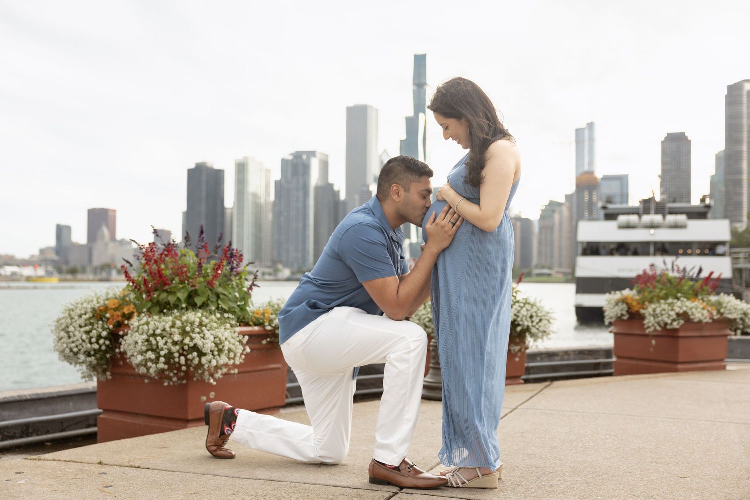 Man kneeling and kissing pregnant woman's belly by Chicago waterfront with city skyline behind them.