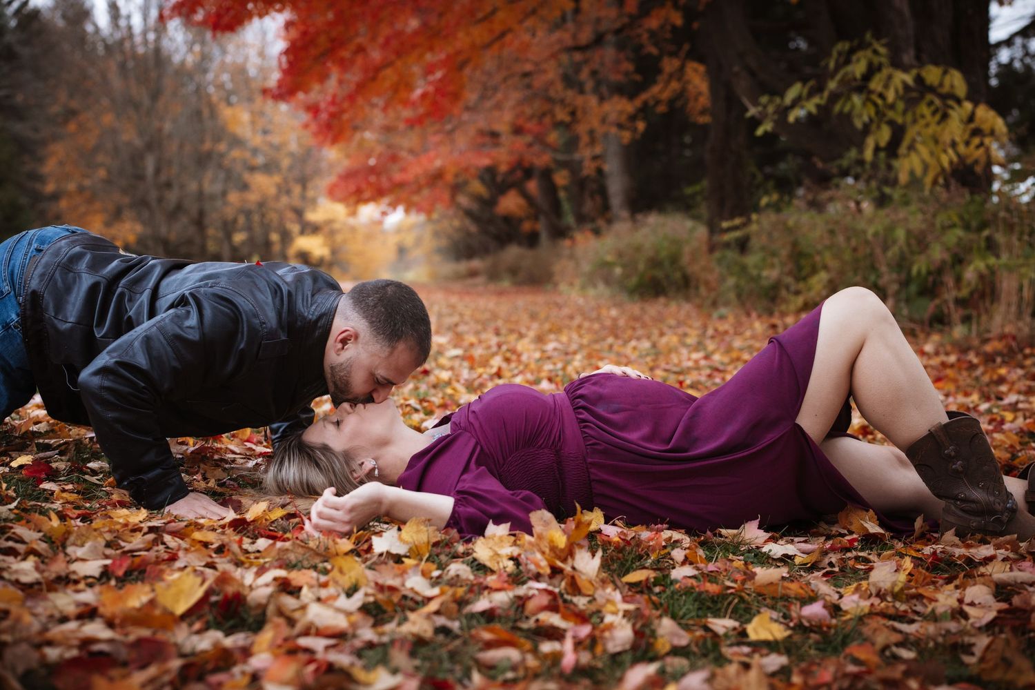 Man leaning down to kiss his pregnant partner lying on colorful autumn leaves in a scenic fall landscape.