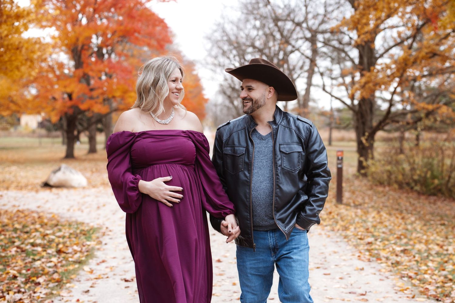 Smiling pregnant couple holding hands and gazing at each other among vibrant fall foliage.