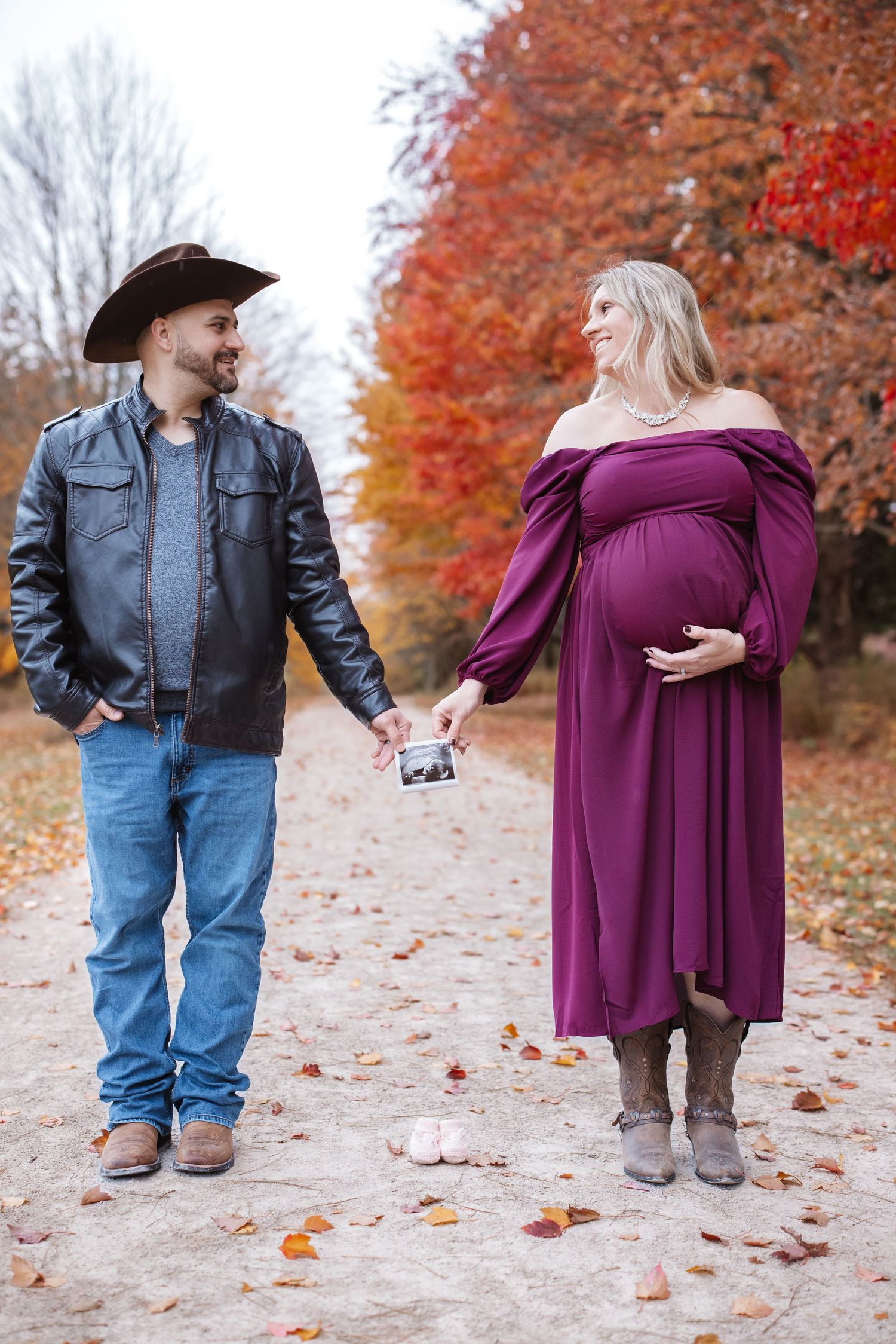 Smiling pregnant couple holding ultrasound photo on colorful fall foliage lined pathway.