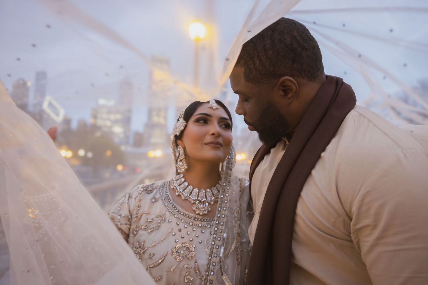 Bride adorned in traditional South Asian bridal jewelry and groom share tender moment under veil.