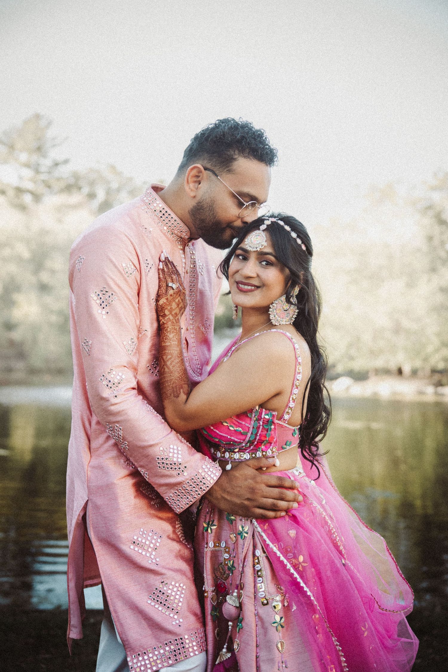 Indian groom embracing bride in pink lehenga near a reflective lake backdrop.