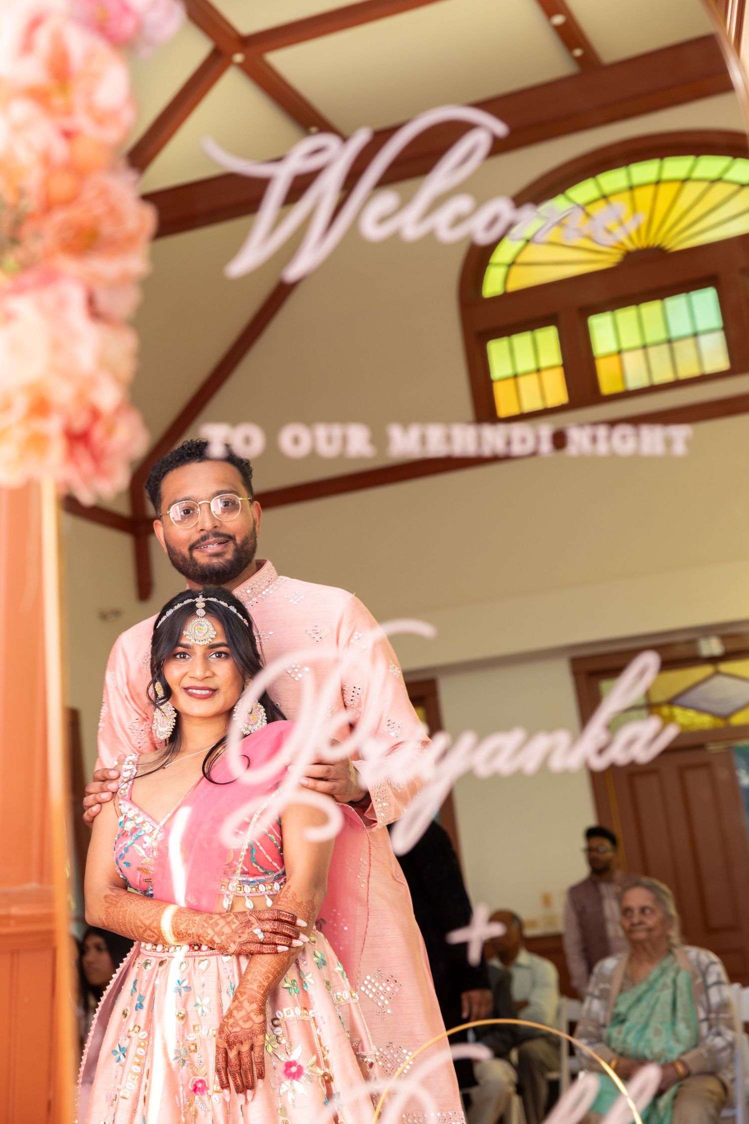 Indian couple posing at mehndi night event with decorative welcome sign, bride in pink floral lehenga.