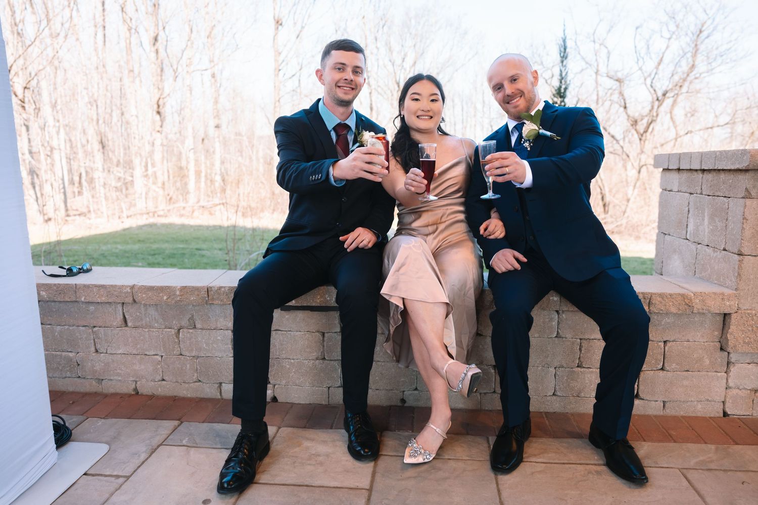 Two men in navy suits and a woman in blush dress toast with champagne glasses at an outdoor wedding.