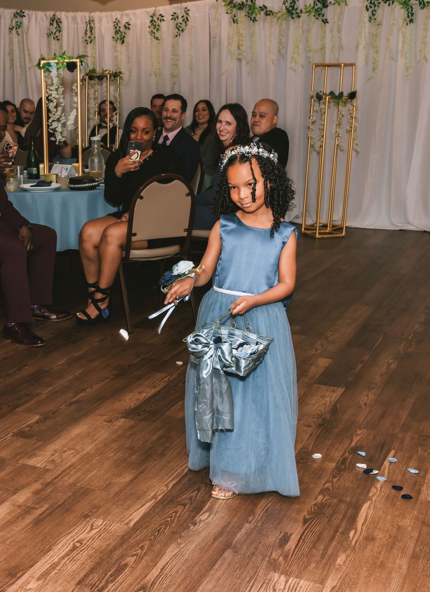 Young flower girl in blue dress walks down aisle scattering petals at elegant indoor wedding ceremony.