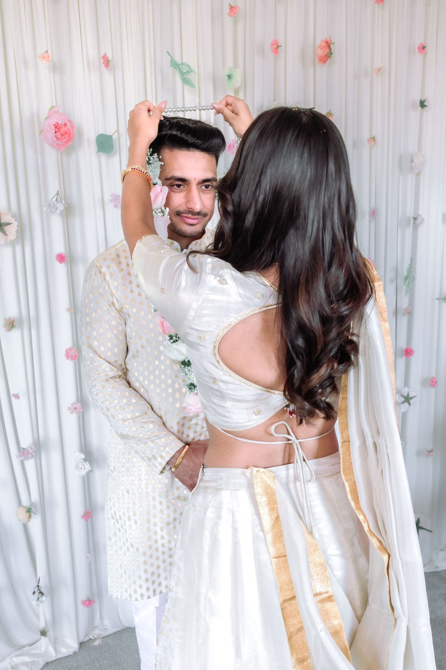 Indian bride and groom embracing and smiling during traditional wedding ceremony with floral backdrop.