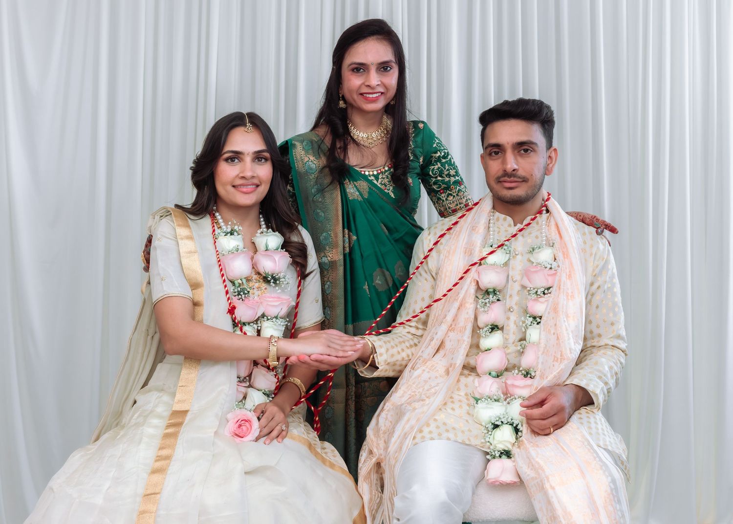 Indian bride and groom in traditional white and cream wedding outfits posing with female guest in green saree.