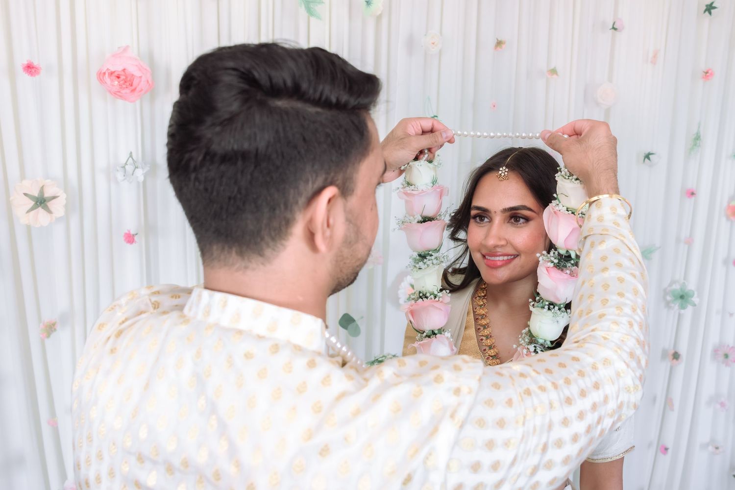 Groom framing brides face with floral garland during Indian wedding ceremony against decorative white backdrop.