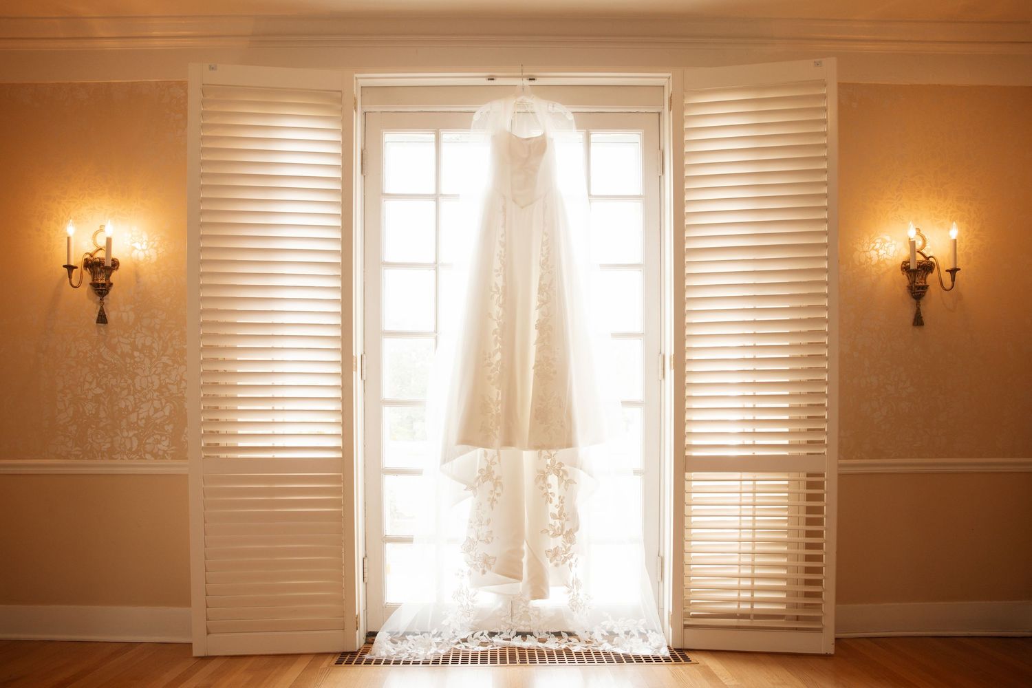 Beautiful lace bridal gown displayed between open white plantation shutters with golden wall sconces flanking the doors.