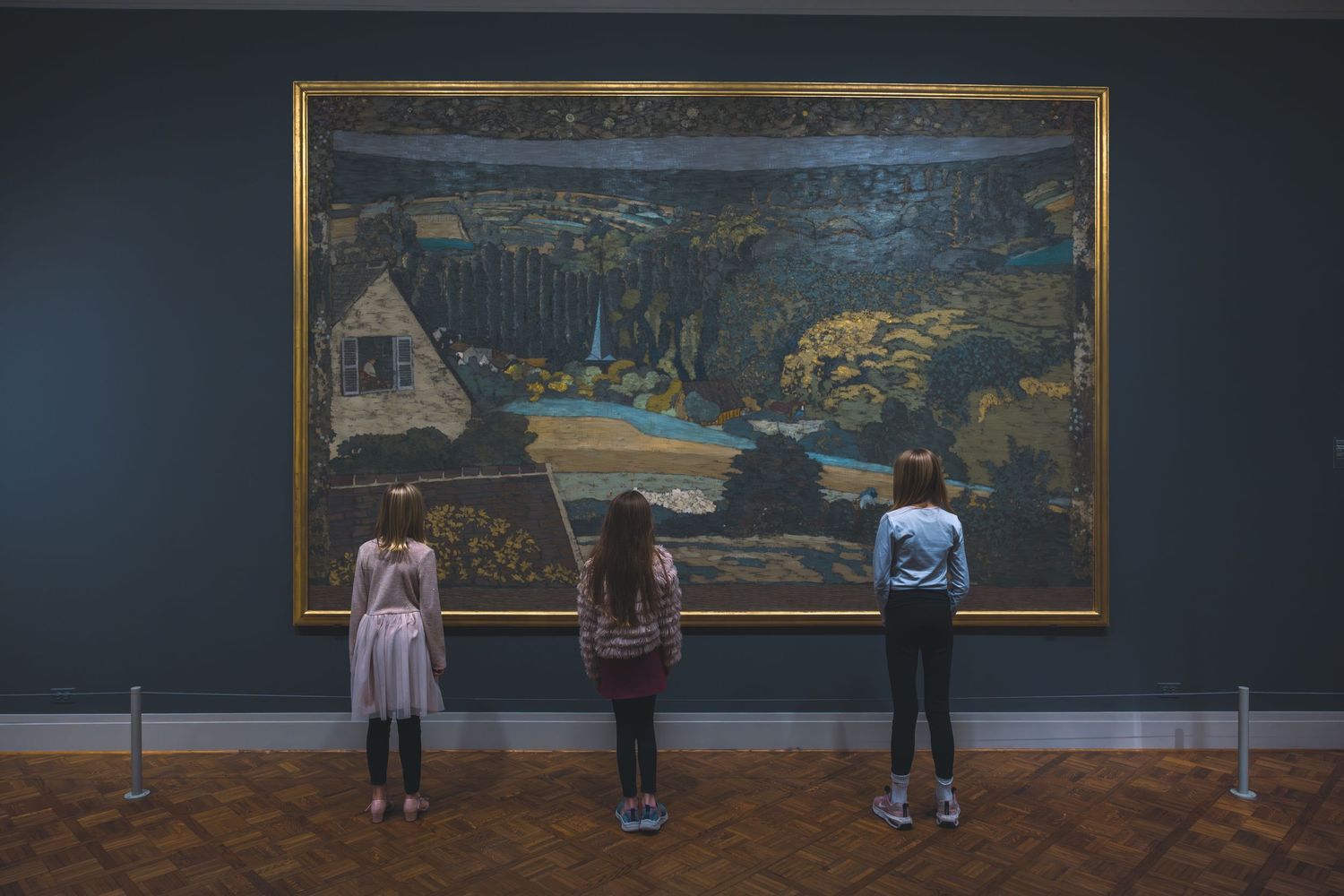 Three children standing before a large landscape painting in a dimly lit art museum.