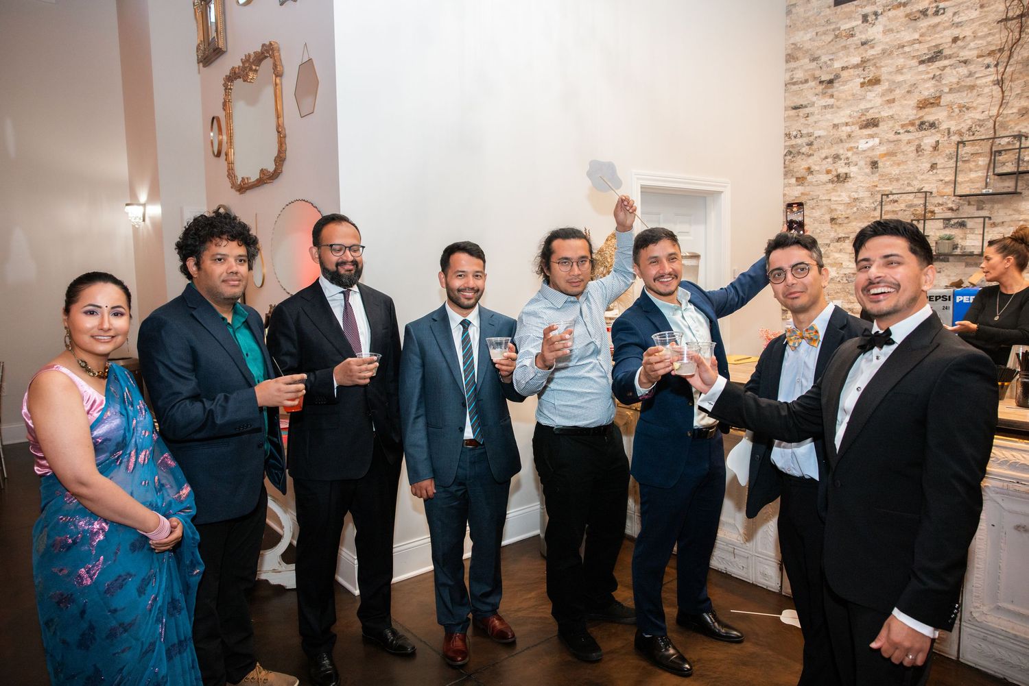 Group of seven men in formal attire toast and celebrate at Indian wedding reception.
