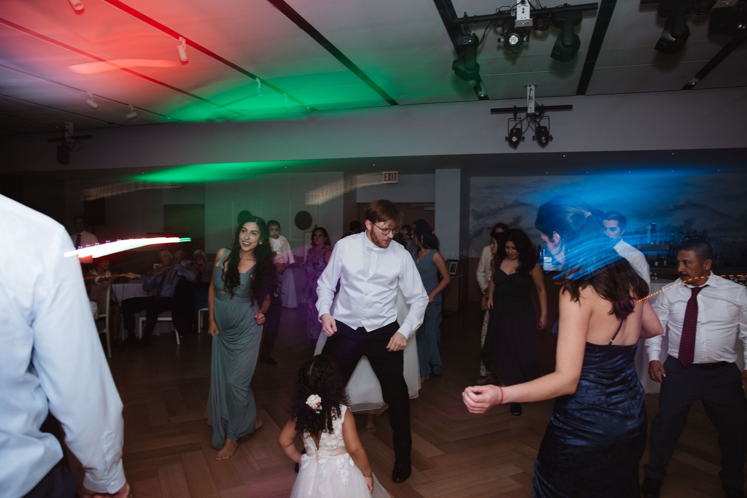Wedding guests dancing energetically on dance floor with colorful red and green stage lighting at reception.