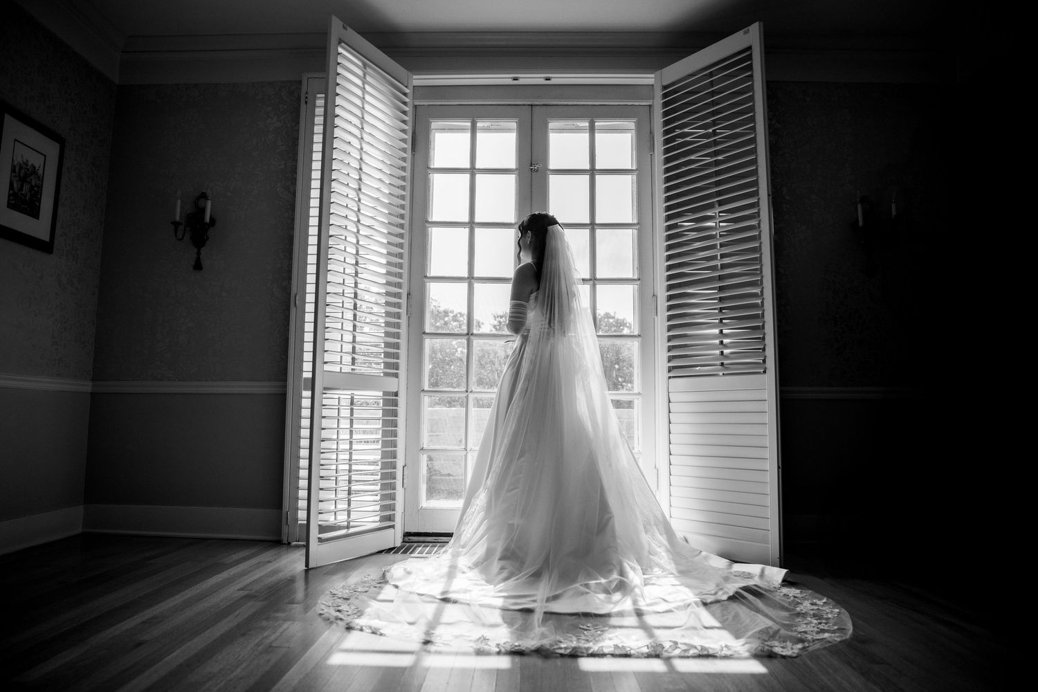 Black and white photo of bride in gown with long veil standing before French doors.