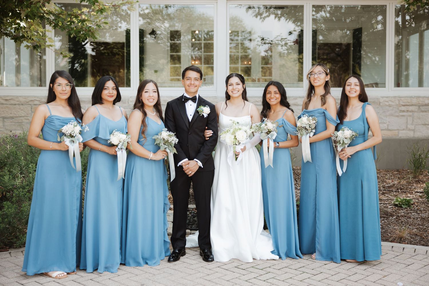 Bride and groom surrounded by six bridesmaids in dusty blue gowns holding white bouquets outdoors.