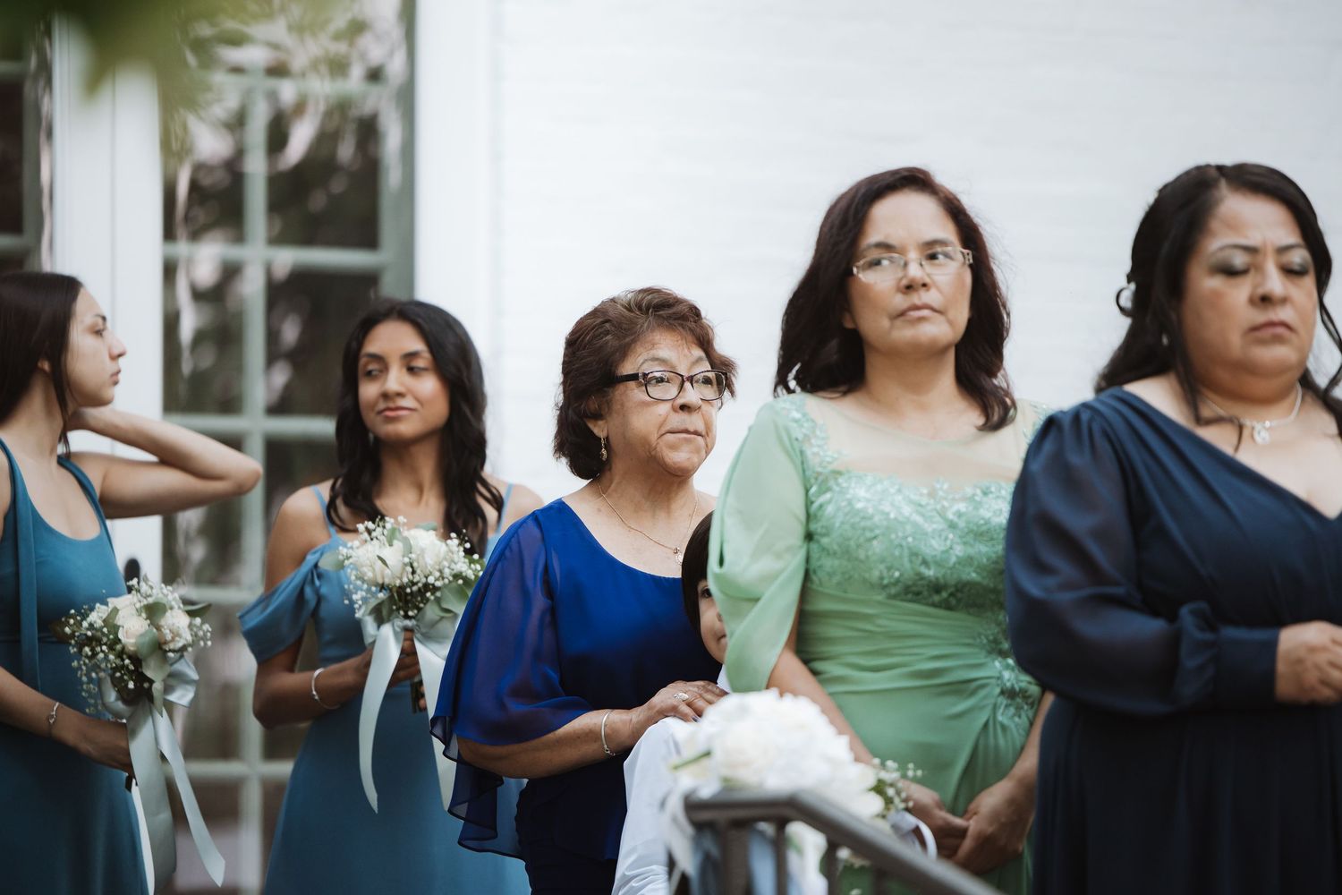 Women in formal attire watch a wedding ceremony outdoors, with bridesmaids holding bouquets.