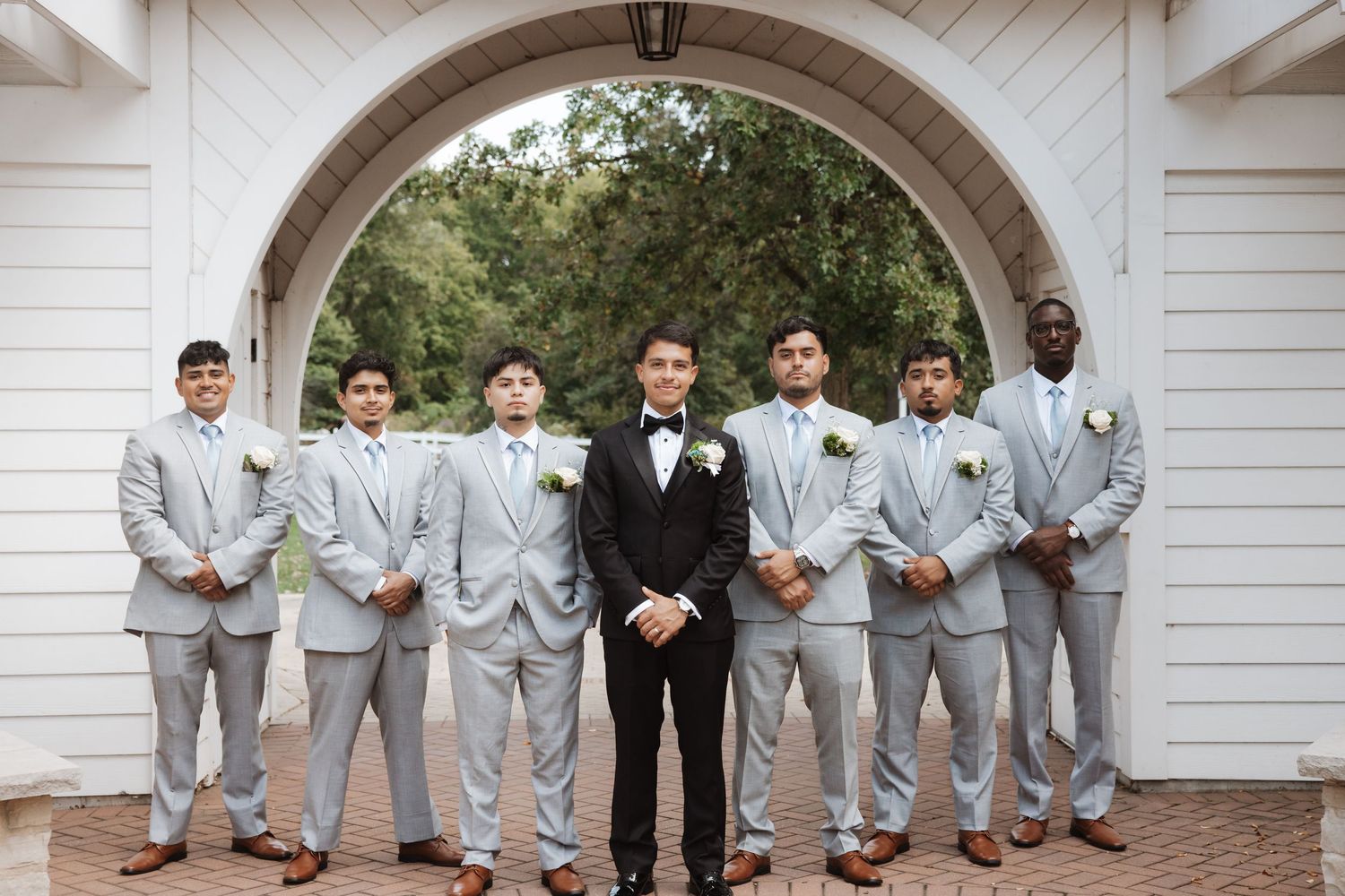 Wedding party poses under white arch with groom in black and groomsmen in gray.