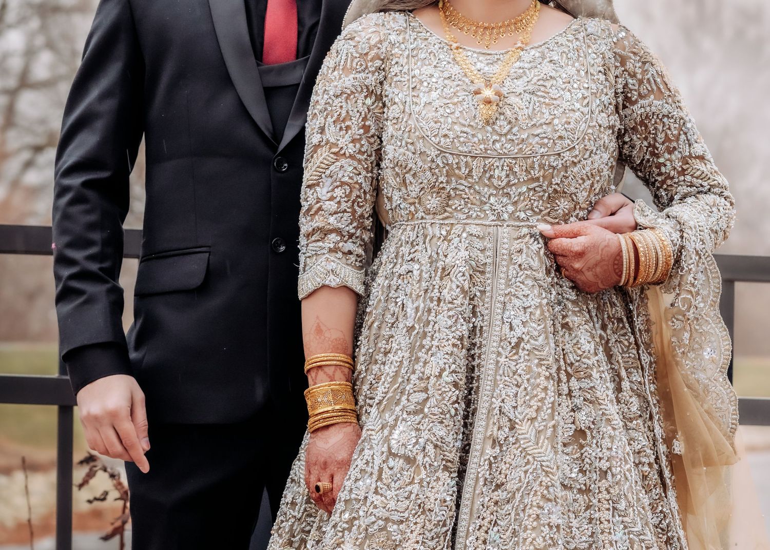 South Asian bride in heavily embroidered gold lehenga holds groom's hand wearing gold bangles and jewelry.