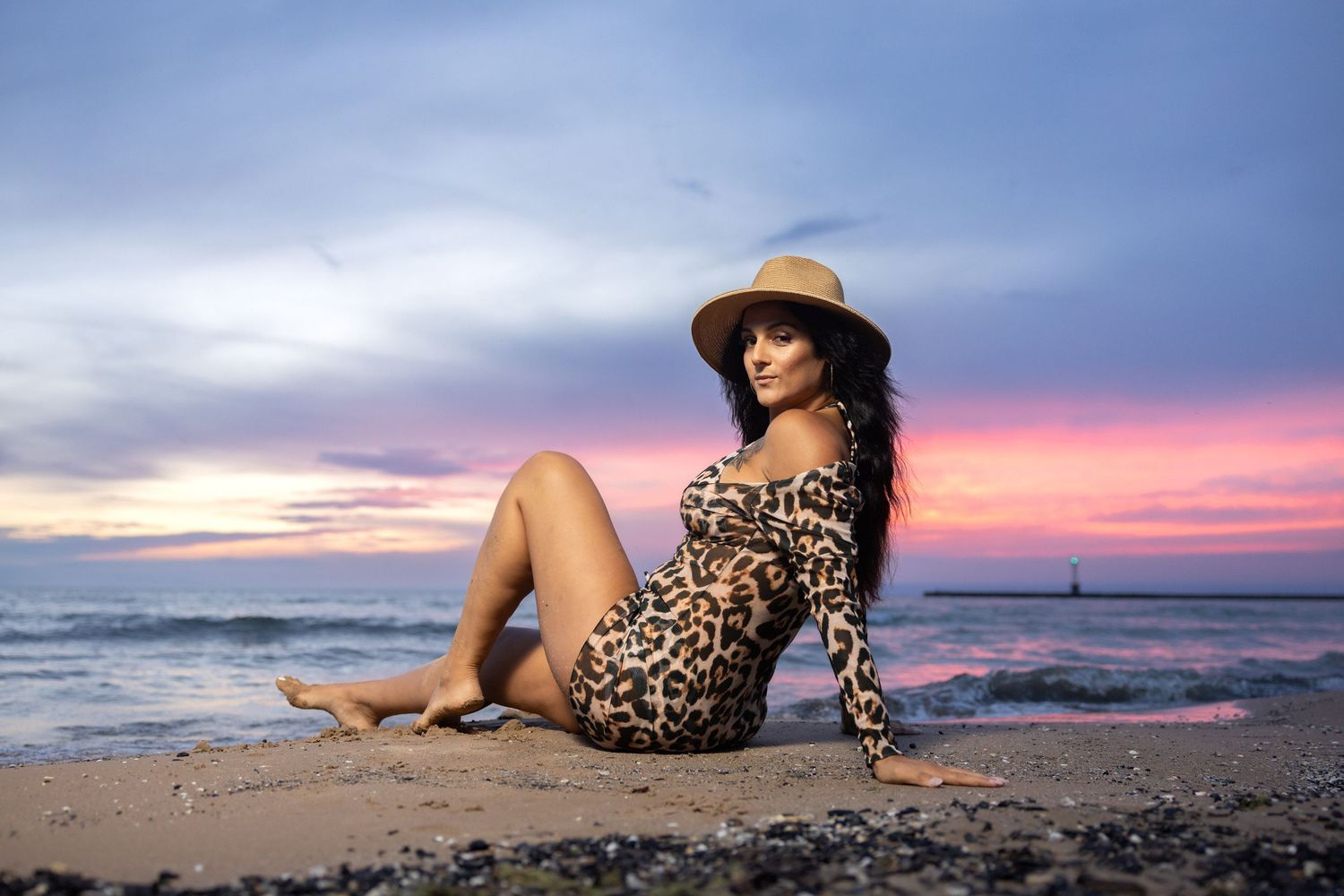 Woman in leopard dress and straw hat sitting on beach at colorful sunset.