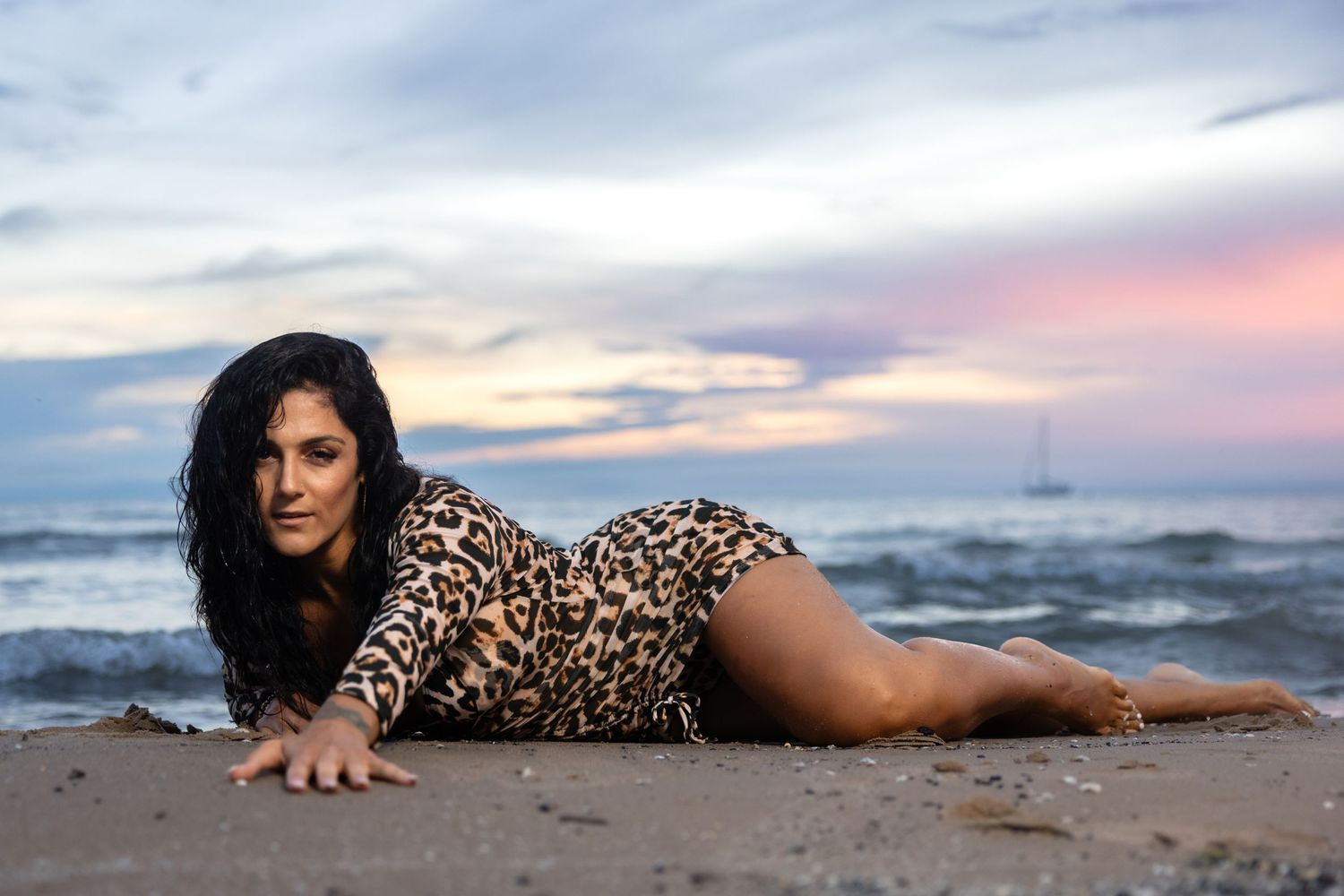 Woman in leopard print dress lying on sandy beach at dusk with ocean waves and pastel sky.