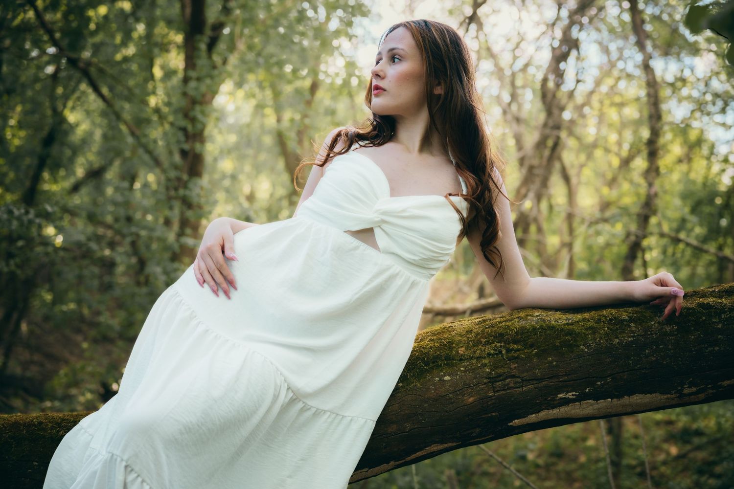 A woman in a flowing white dress leans against a mossy log in a dreamy, softly lit forest setting.