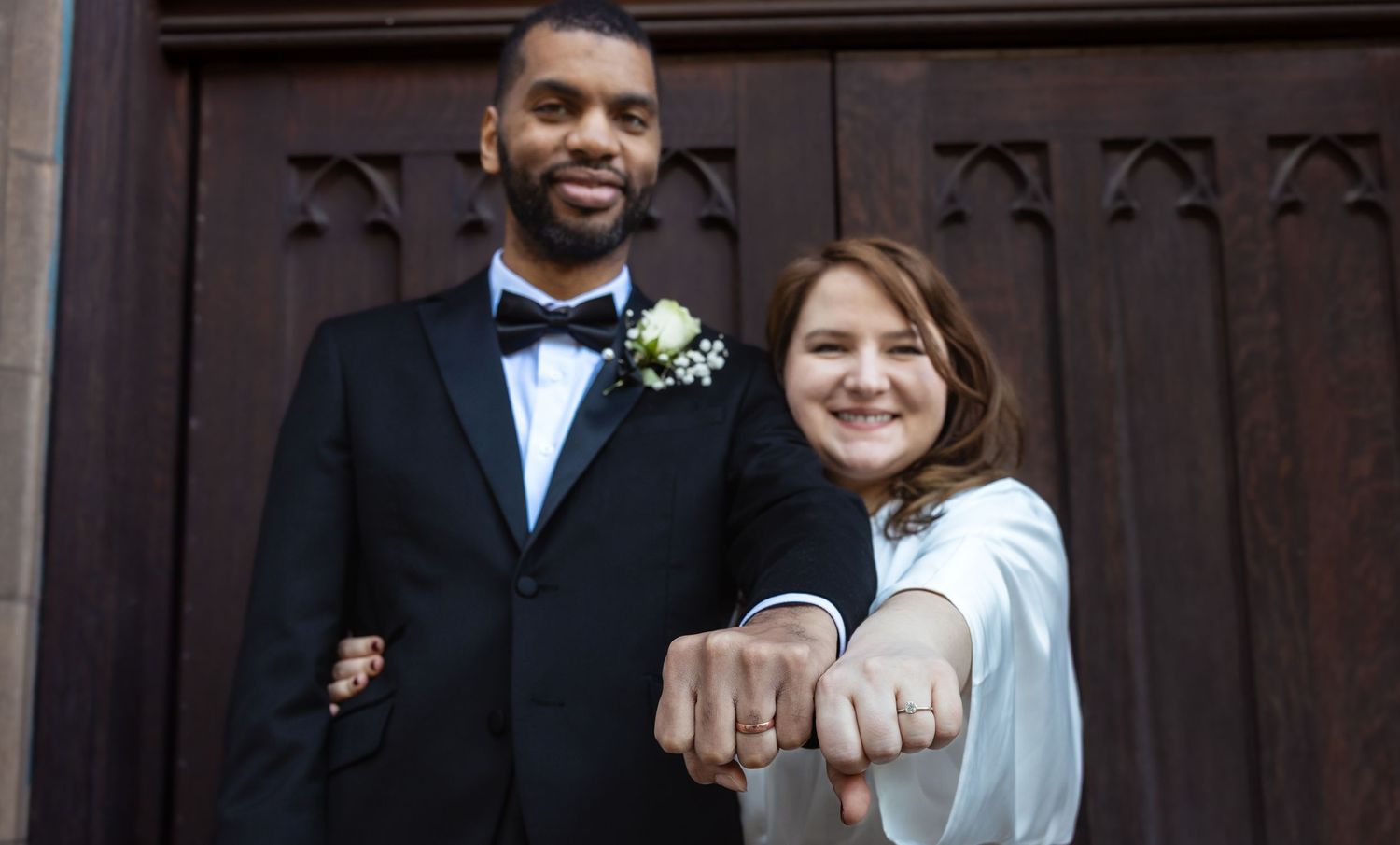Newlywed interracial couple smiling and showing wedding rings outside a church.