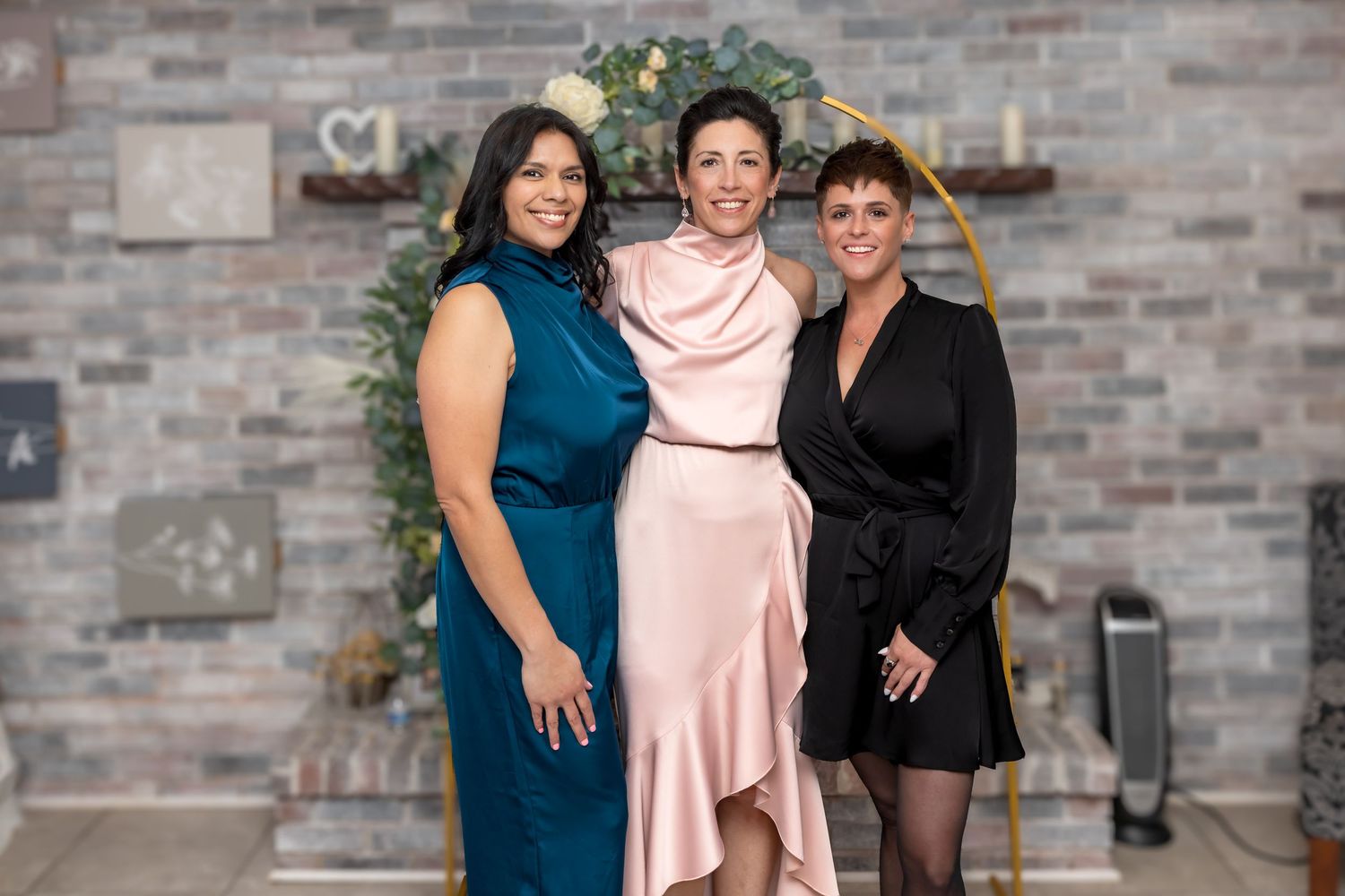 Three women in elegant formal dresses posing together at an event with stone wall backdrop.