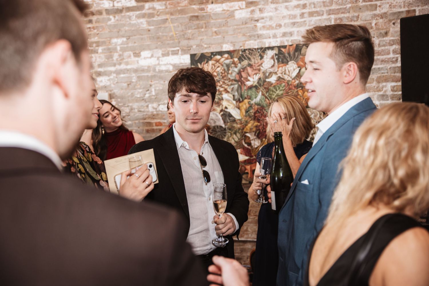 Guests mingle at an elegant indoor cocktail party near a brick wall with artwork.