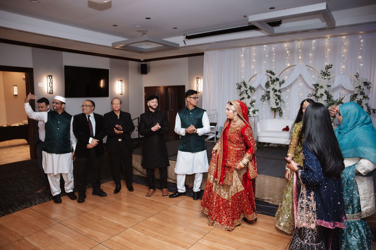 South Asian wedding guests in traditional attire gather around bride and groom in elegant venue.