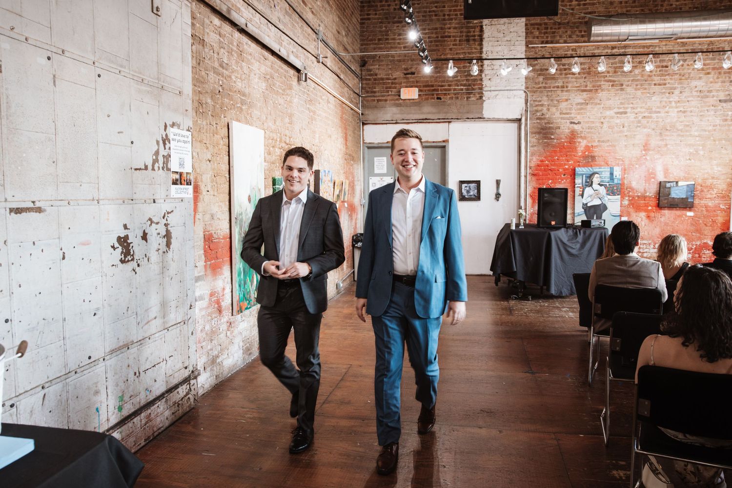 Two well-dressed men walk confidently through an industrial-style event venue.