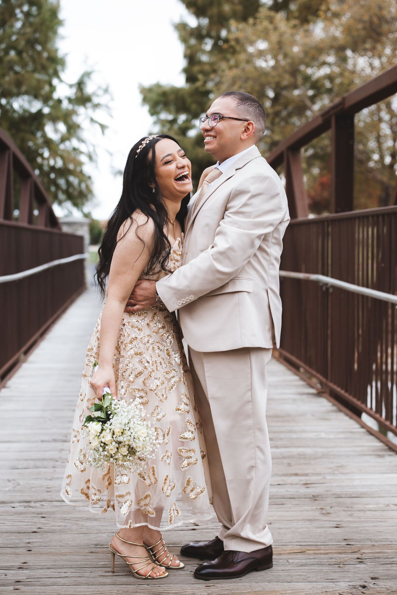 Laughing newlywed couple embracing on wooden bridge, bride in gold embroidered dress.