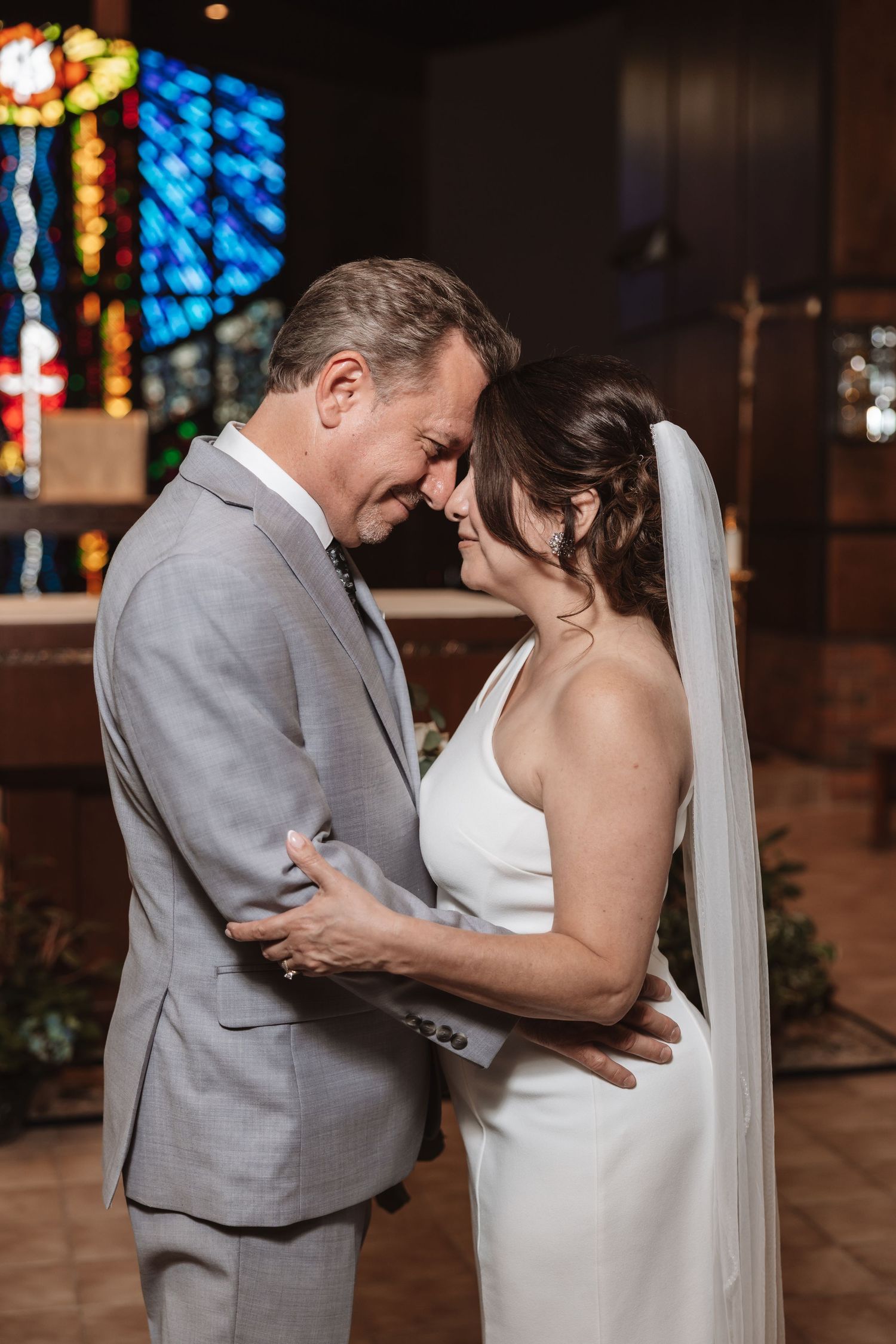 Bride and groom share an intimate forehead touch in a church, dressed in white gown with veil and gray suit.