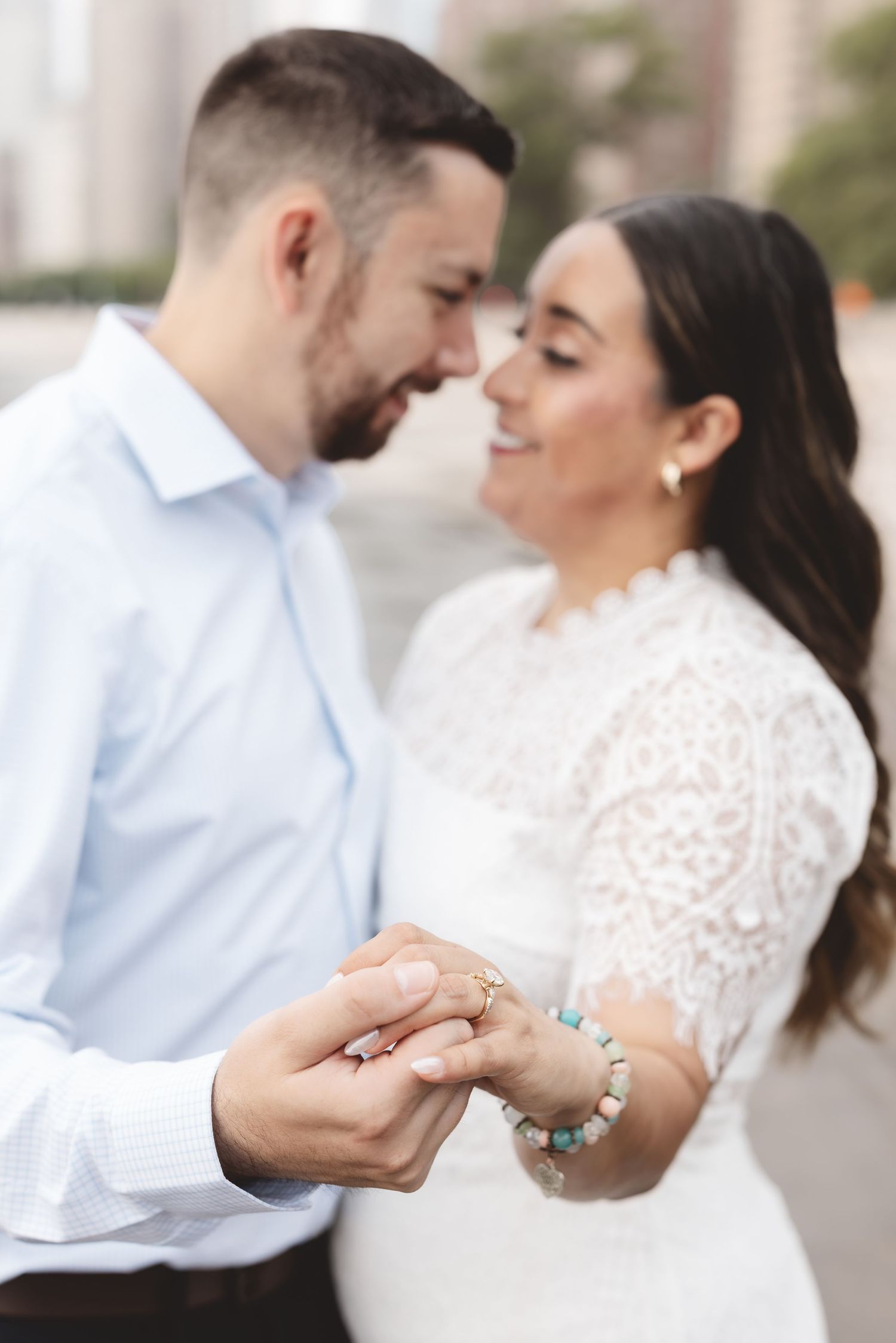 Engaged couple smiling face-to-face while holding hands, showcasing engagement ring and turquoise bracelet.