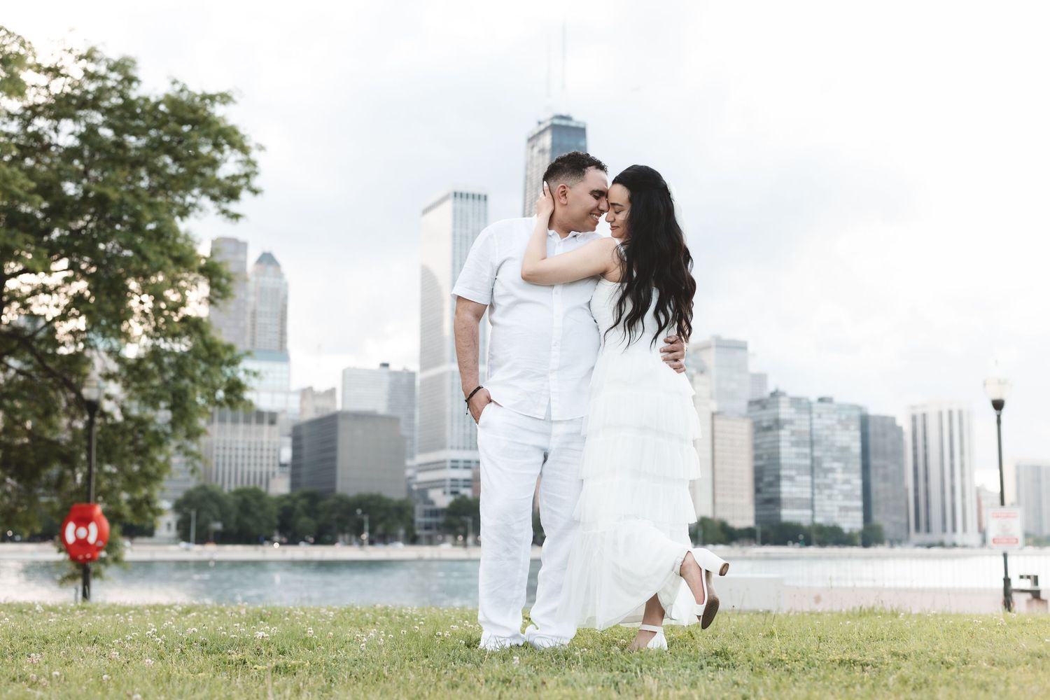 Couple in white outfits embracing on grass with Chicago skyline and lake in background.