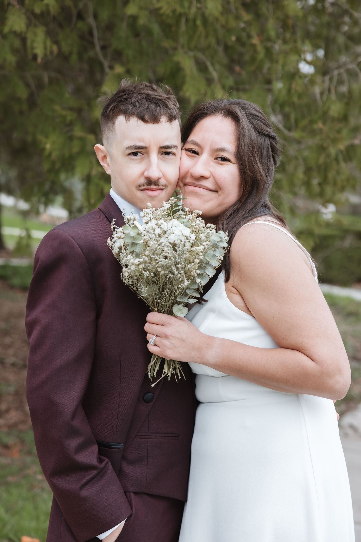 Bride in white dress holding white bouquet embraces groom in burgundy suit outdoors at wedding portrait.