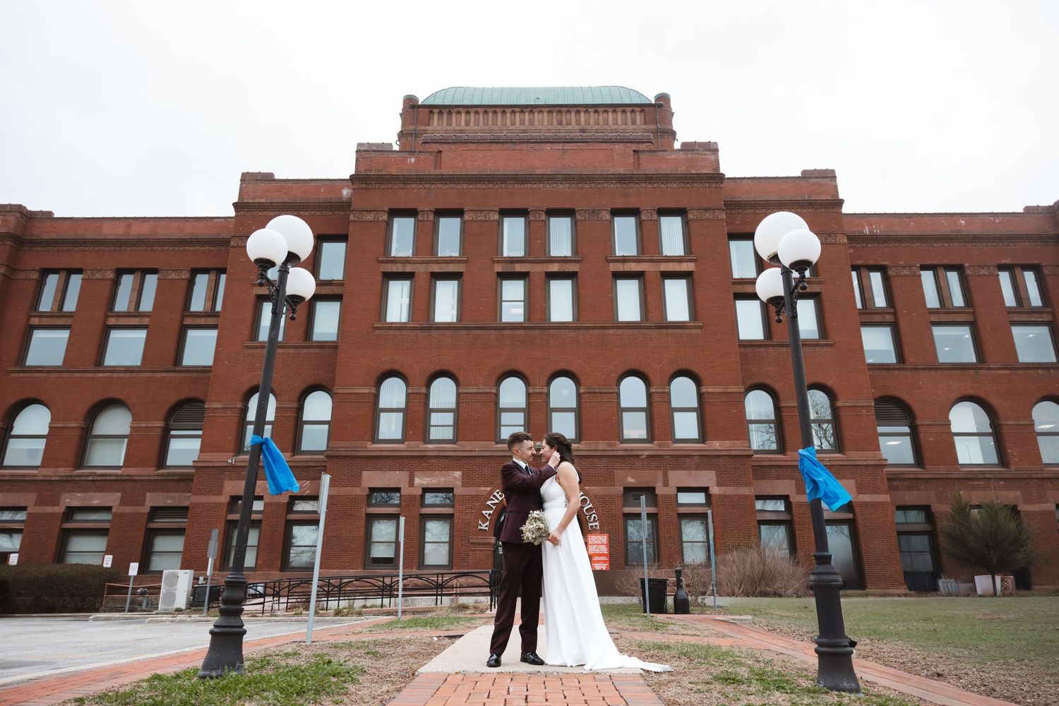 Bride and groom embrace in front of a historic red brick building on their wedding day.