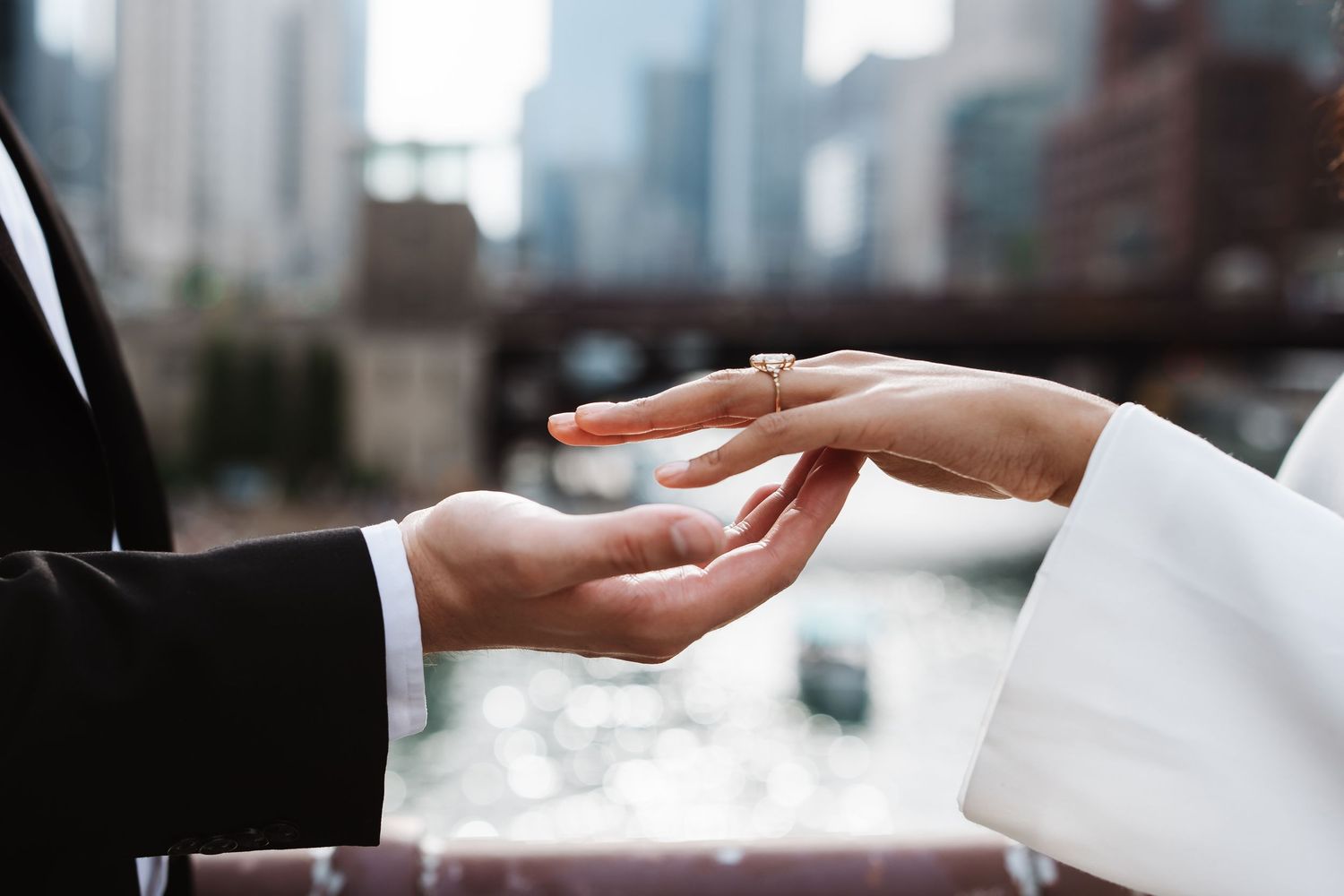 Close-up of groom placing diamond engagement ring on bride's hand with city river backdrop.