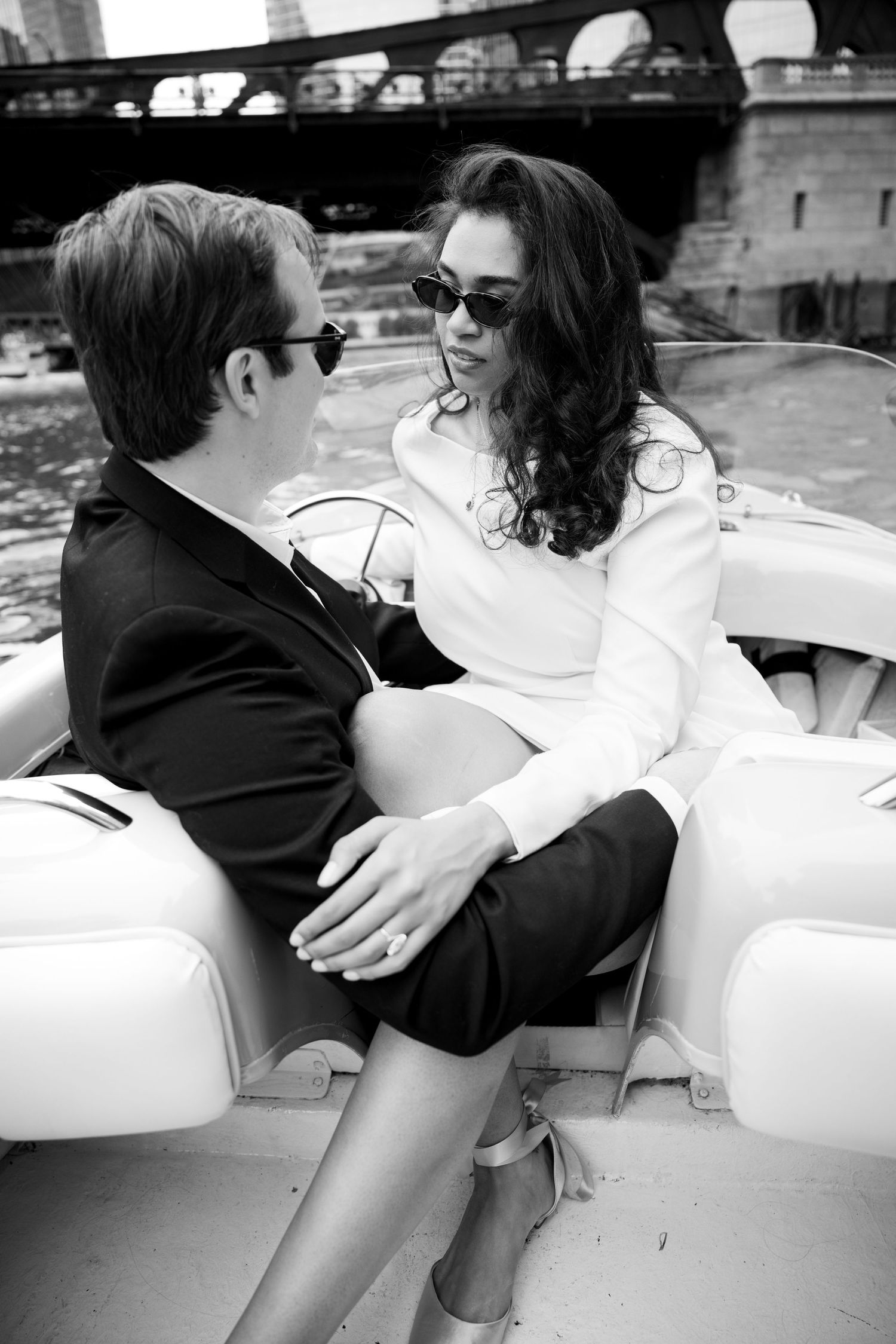 Couple in formal attire sitting intimately on a boat, wearing sunglasses, black and white photo.