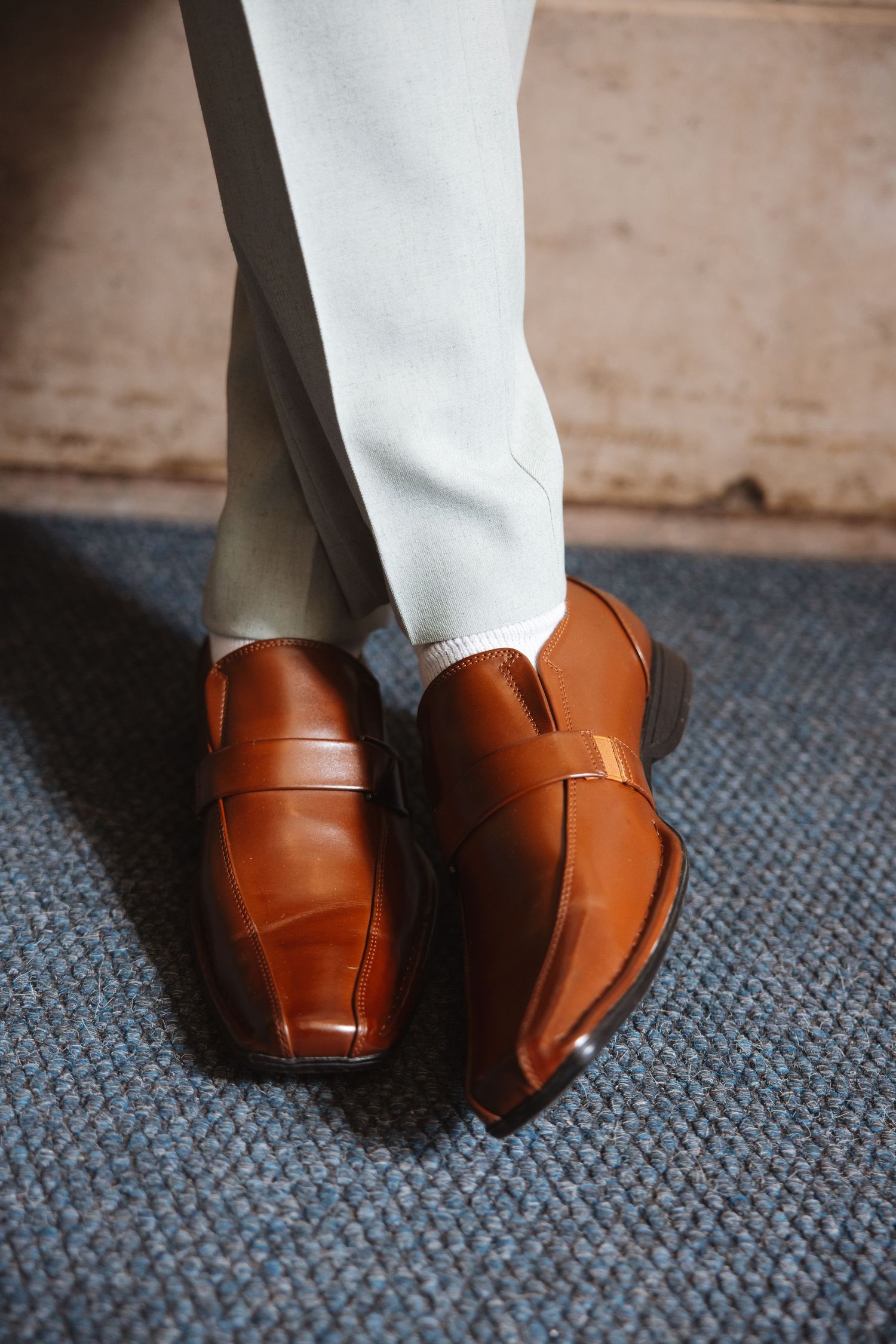 Close-up of groom wearing tan brown leather loafers with white socks and light gray dress pants on blue carpet.