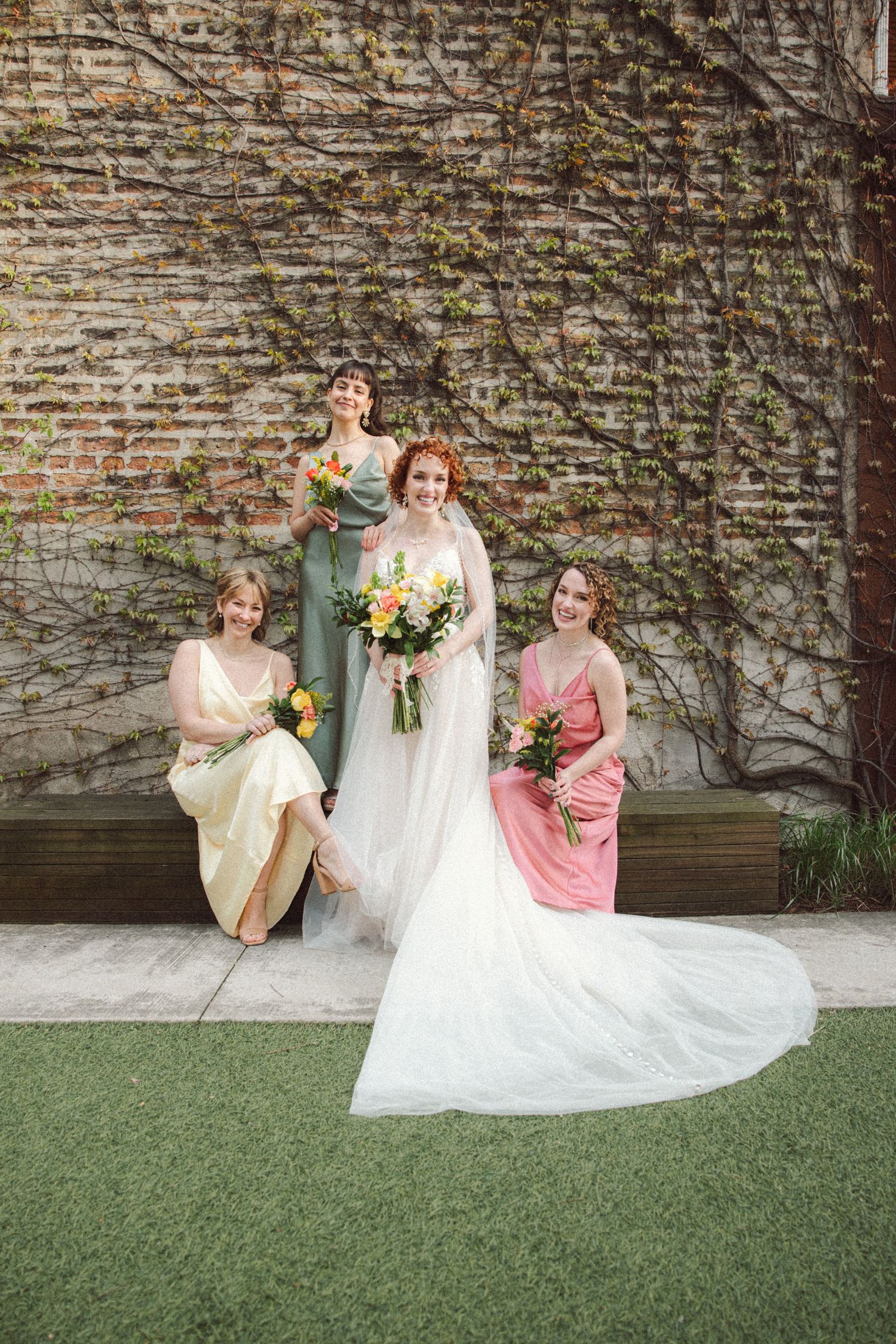 Bride in white gown with three bridesmaids in colorful dresses posing against ivy-covered brick wall.