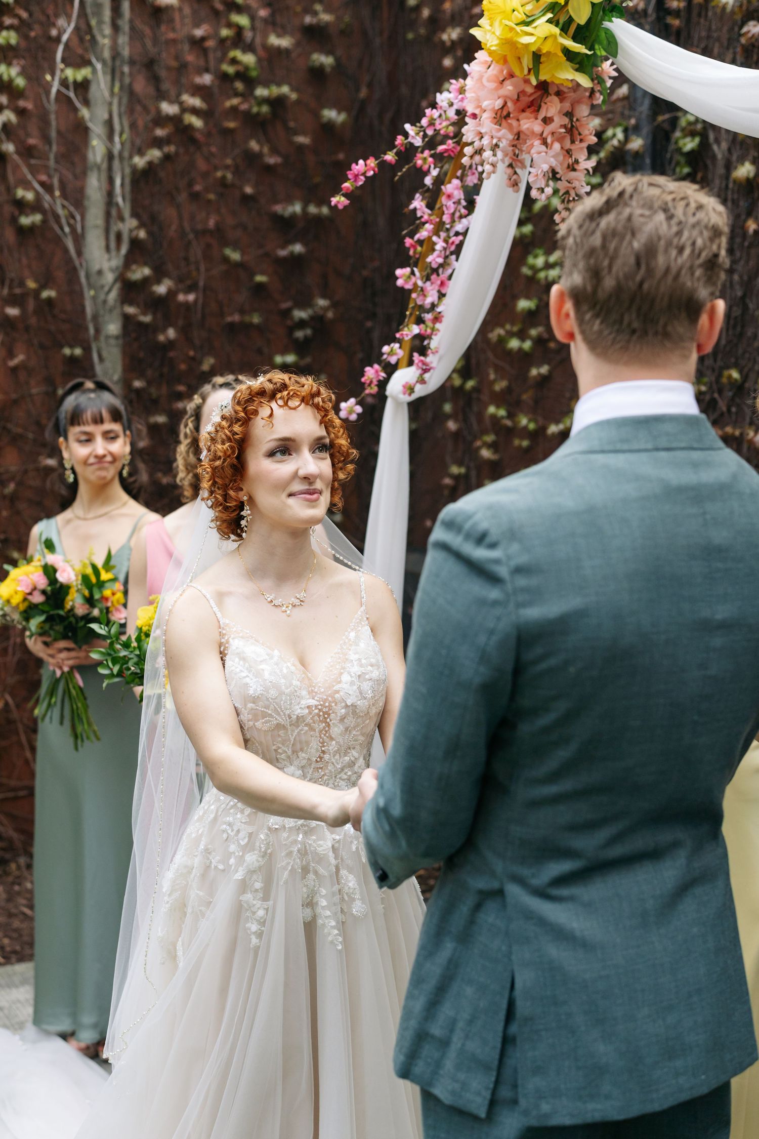 Bride with curly red hair in lace gown holds groom's hands at outdoor wedding ceremony under floral arch.