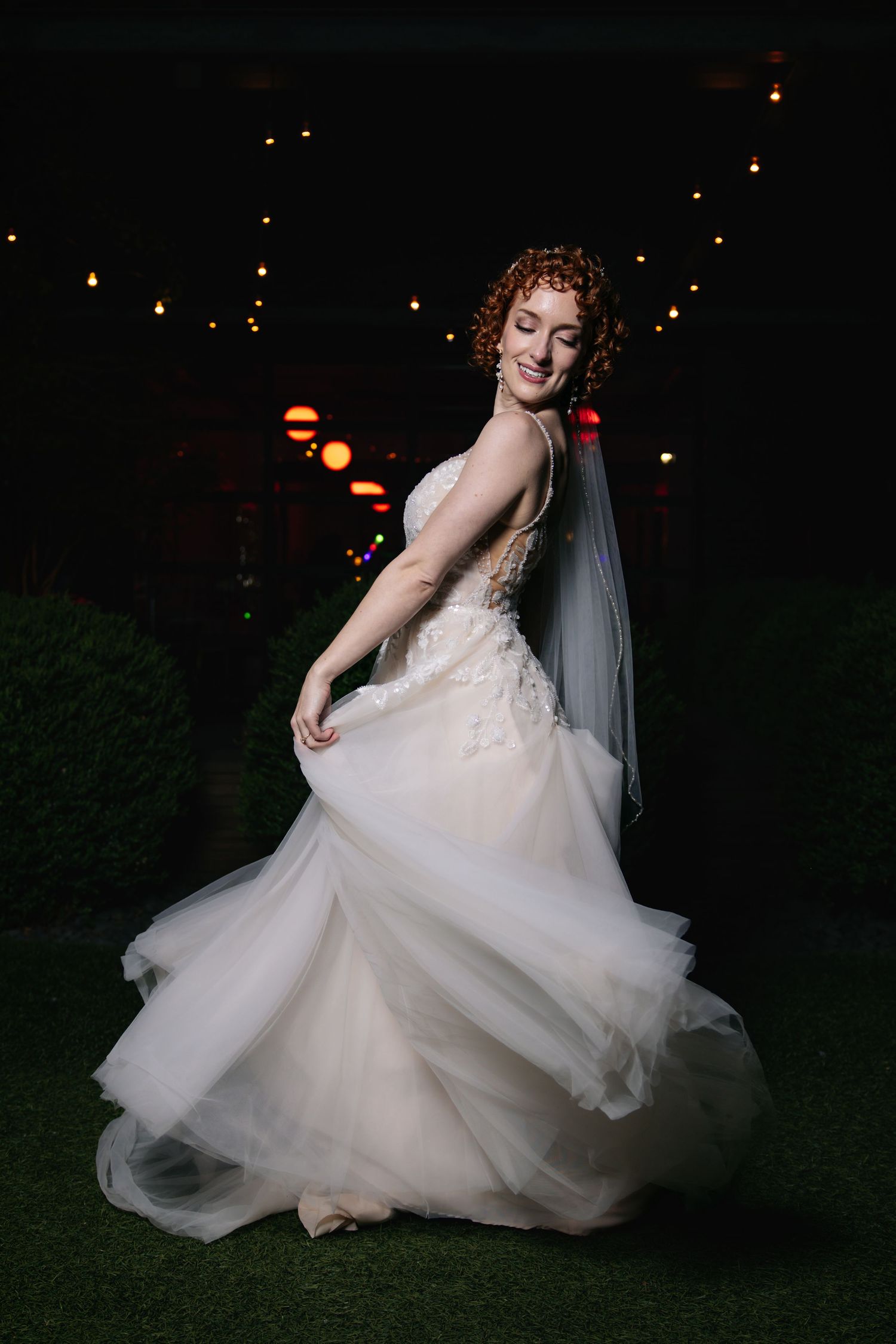 Curly red-haired bride twirls in white tulle wedding gown under string lights in dark outdoor setting.
