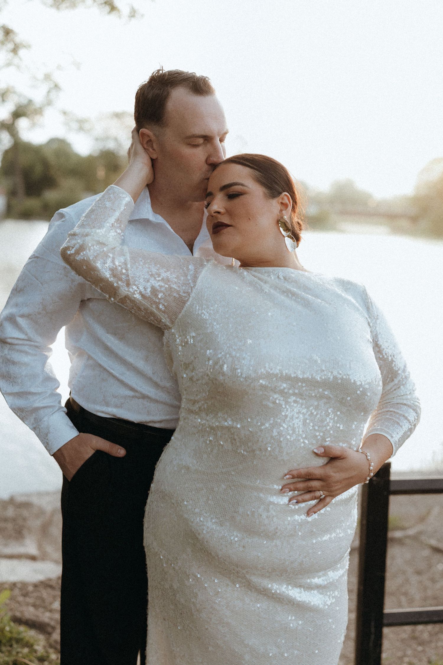 Groom tenderly kissing bride wearing elegant sequined white dress near a beautiful waterfront setting.